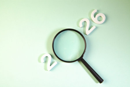 Magnifying glass and white wooden numbers 2026 on a green background.