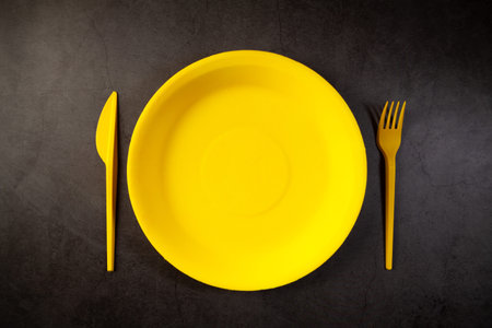 Empty yellow plate, fork and knife on black background. Top view