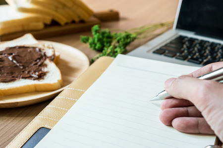 A man holding pen on top of the notebook. The working table include laptop, notebook and breakfast with sliced bread with chocolate spread. Focused on hand.の写真素材