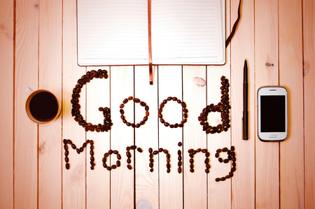 Good Morning inscription made with coffee beans between white cell phone, cup of coffee, pen and notebook on wooden table. Coffee in the morning. Ready to work.の写真素材