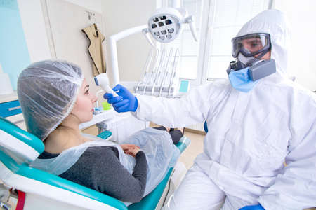 Doctor in protective suit uniform and mask checking temperature of young female patient. Coronavirus outbreak. Covid-19 concept.の写真素材