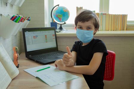 Distance learning. Schoolboy in a medical mask at home is doing homework on a laptop. School books...