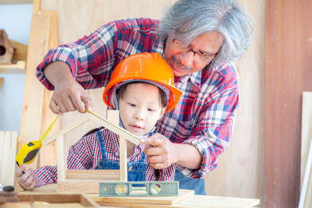 Asian senior carpenter man teaching asian little girl craftswoman measuring size of wooden house toy in carpentry shop.の写真素材
