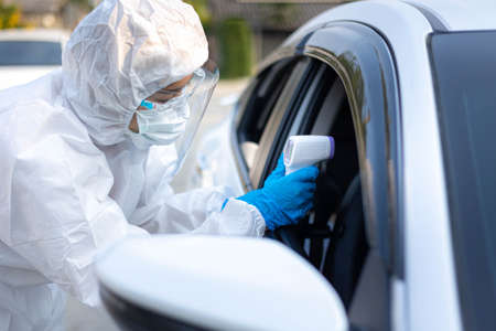 Medical staff with PPE use infrared thermometer gun to check body temperature at drive thru station...