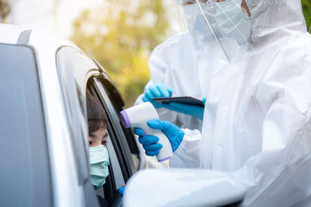 Medical staff with PPE use infrared thermometer gun to check body temperature and take notes on the tablet at drive thru station in hospital. New normal healthcare and medical concept.の写真素材