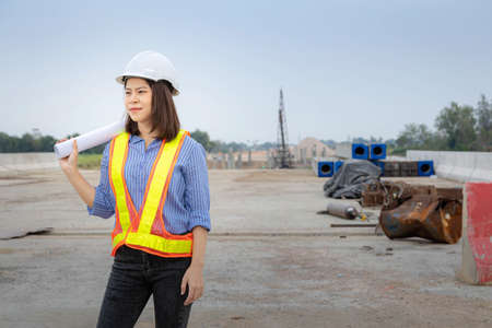 A female architect leader standing at the bridge construction,  Wearing a safety helmet and holding the blueprint of the project.の写真素材