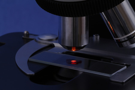 The drop of a red liquid like a blood illuminated on a slide of a dark microscopeの写真素材