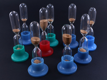 Group of hourglasses of different colors standing on dark surface. One red sand clock near the central area of group. The top view. Focus on front raw of hourglass two front blue sandglass.の写真素材