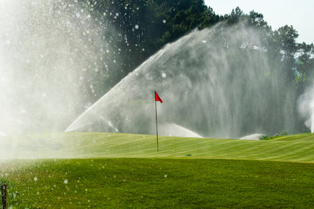 golf course with water sprinkler and green grass and red flagの写真素材