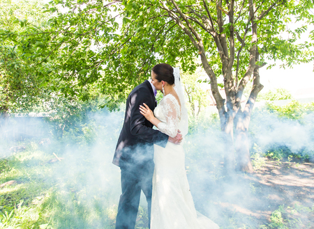 couple in wedding attire  with a bouquet of flowers and greenery is in the hands kissing against the backdrop of the misty gardenの写真素材
