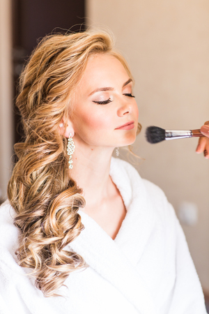Wedding makeup artist making a make up for bride. Beautiful sexy model girl indoors. Beauty blonde woman with curly hair. Female portrait. Bridal morning of a cute lady. Close-up hands near faceの写真素材