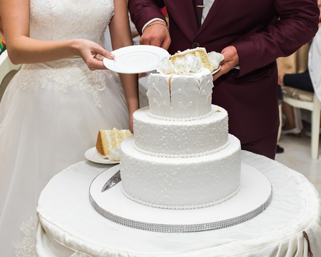 Beauty bride and handsome groom are cutting a wedding cake. Couple in the restaurant with colorful pie. Beautiful model girl in white dress. Man in suit. Female and male portrait. Cute lady and guyの写真素材