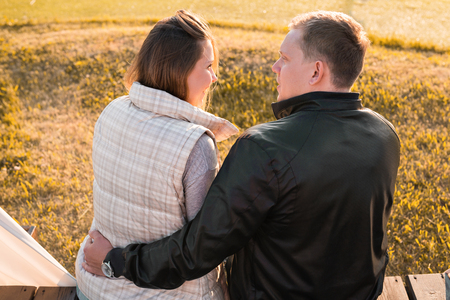 Romantic young couple enjoying a date sitting in a close embrace on a park bench overlooking a lake, view from behindの写真素材