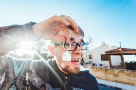 Happy funny man holds house keys on house shaped keychain in front of a new homeの写真素材