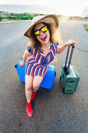 Happy traveler woman sitting on a suitcase on the road and smiling. Concept of travel, journey, tripの写真素材