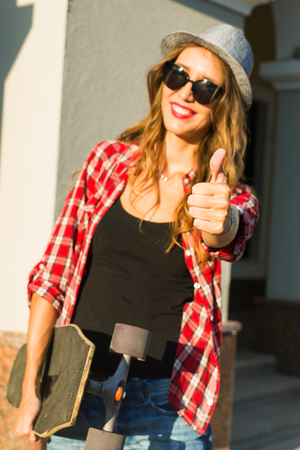 summer holidays, extreme sport and people concept - happy girl showing thumbs up gesture with skateboard on city streetの写真素材