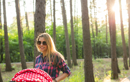 family, child and parenthood concept - Young mother walking and pushing a stroller in the park with newborn. Beautiful happy mother with pram outdoors. Summer walks in the sunny day. Girl with carriage.の写真素材