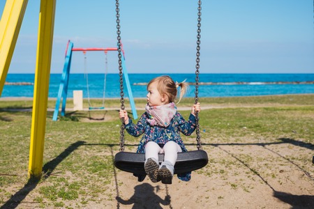 Little baby girl fly with fun on rope swing on sea beach with waves and surf in tropical island. Baby, people activity on summer family vacation with child.の写真素材