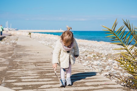 Child, childhood and people concept - Baby girl sits on square road and play with stick near sea...