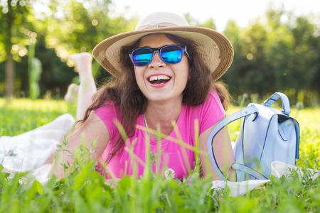 portrait of happy young woman with hat and sunglasses lying in park on picnicの写真素材