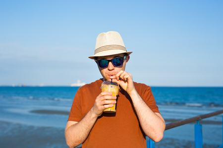 Portrait of cheerful attractive bearded hipster young man drinking iced coffee or frappe on the background of the seaの写真素材
