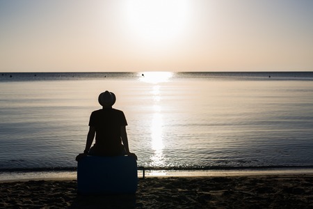 Back view silhouette of man sitting on suitcase on blue sea outdoors background. Arriving at destination.の写真素材
