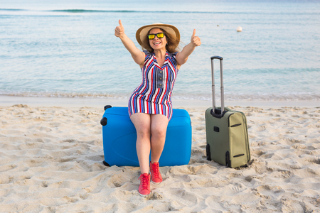 Beautiful young lady with a blue suitcase showing thumbs up gesture on the beach. People, travel, vacation and summer conceptの写真素材