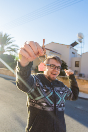 Young funny man holding house keys on house shaped keychain in front of a new home.の写真素材