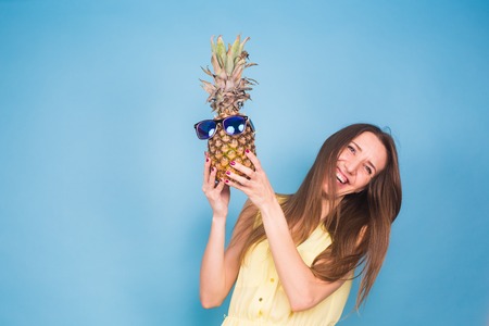 Happy young woman holding a pineapple with sunglasses on a blue backgroundの写真素材