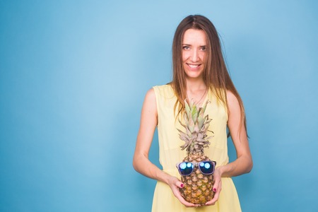 Happy young woman holding a pineapple with sunglasses on a blue backgroundの写真素材