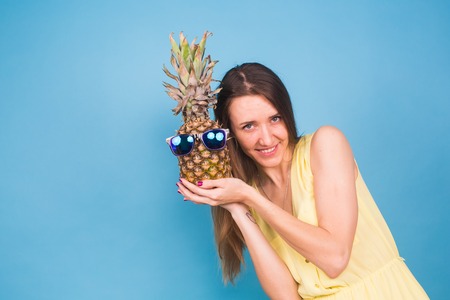 Happy young woman holding a pineapple with sunglasses on a blue backgroundの写真素材