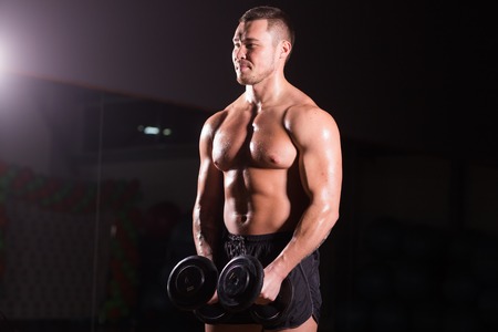 Muscular young man lifting weights on dark backgroundの写真素材