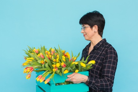 Happy middle-aged woman florist wearing glasses with box of tulips on blue backgroundの写真素材