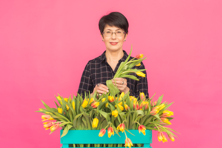 Floristics, holidays and people concept - Middle-aged woman holding box of tulips on a pink backgroundの写真素材