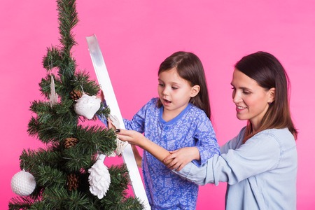 Holidays, family and christmas concept - pretty girl with mum are decorating a christmas tree on pink backgroundの写真素材