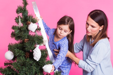 Christmas, childhood and people concept - child girl standing on step-ladder decorating the christmas tree with her mother on pink backgroundの写真素材