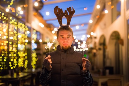 Christmas, people and winter holidays concept - surprised man in horns of deer standing at night street with bengal lights in his handsの写真素材