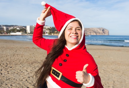 Christmas and holiday concept - Happy woman in Santa Claus costume showing thumbs up on sandy beach backgroundの写真素材