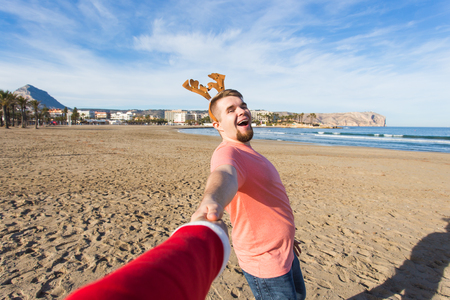 Follow me, holidays and Christmas concept - Happy young man with deer horns holding Santas hand on the beach.の写真素材