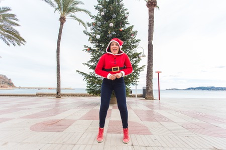 Christmas, holidays and humor concept - young happy woman have fun in christmas suit at summer near palms and christmas treeの写真素材