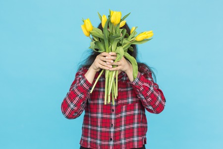 Holidays, flowers and people concept - Funny girl in plaid shirt with bouquet tulips in hands posing on a blue backgroundの写真素材