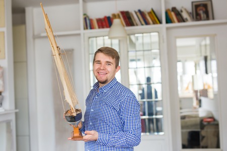 Hobby, interior and collecting concept - handsome man holding the layout of a sailboat in the room