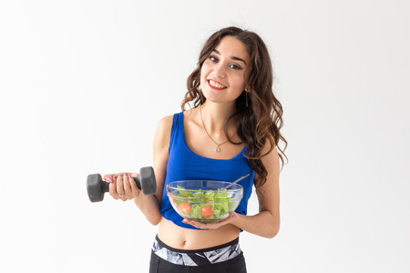 Sport, healthy lifestyle, people concept - young brunette woman with salad and a dumbbell. She is...