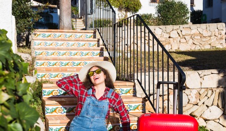 Travel, tourism, emotions and people concept - happy young woman sitting on stairs in a hat with red suitcaseの写真素材