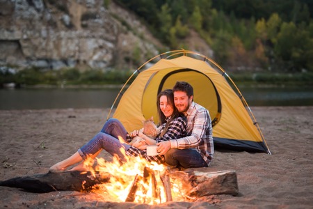 People, tourism and nature concept - Romantic evening, man embrace woman sitting near a campfireの写真素材