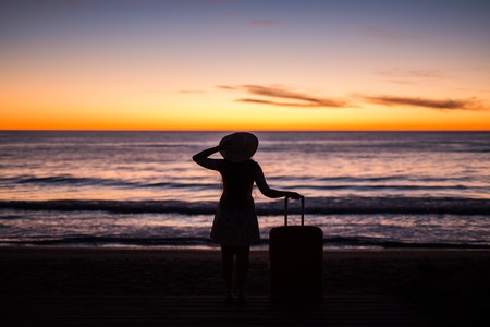 Travel, vacation and summer trip concept - silhouette of young woman in summer dress and hat looking to a seaの写真素材