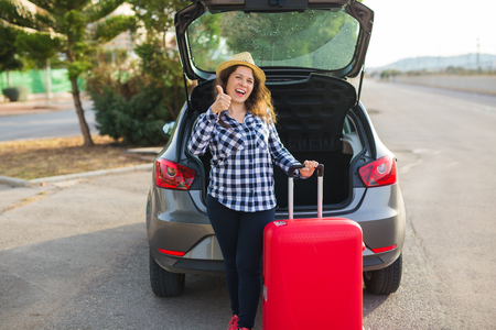 Travelling, holidays and people concept - young woman standing near car in hat with red suitcase. She is ready to go to tripの写真素材
