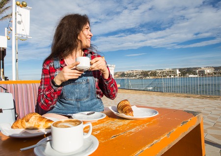 Young woman having breakfast with croissant and coffee at the cafe on the street in front of seaの写真素材