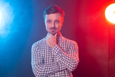 Carming young smiling hipster man is posing against the background of red and blue studio lighting....