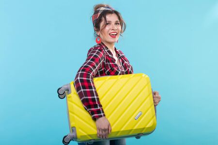 Positive young smiling woman in a plaid shirt holding a yellow suitcase posing on a blue background. Concept of tourism and travelの写真素材